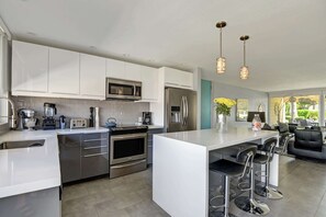A modern kitchen with stainless steel appliances, a central island with bar stools, and a dining area with a view. - Modern Resort Retreat with Mountain Views & Pool (Palm Desert)