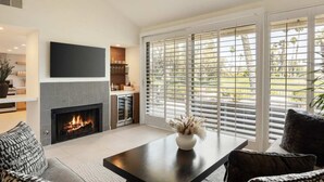A modern living room with a fireplace, a flat-screen TV, and a large window with blinds. - Resort Style Condo Getaway in Monterey Country Club (Palm Desert)