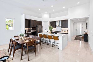 A modern kitchen with a central island, dark wooden cabinets, and a dining area with a table and chairs. - 4 Bedroom Luxury Estate With Pool, Spa, & Firepit (Bermuda Dunes)
