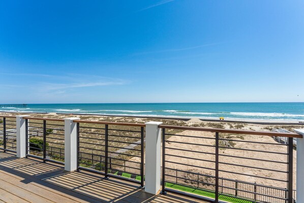Vista de la playa desde una terraza de madera con barandal metálico, playa arenosa y océano.