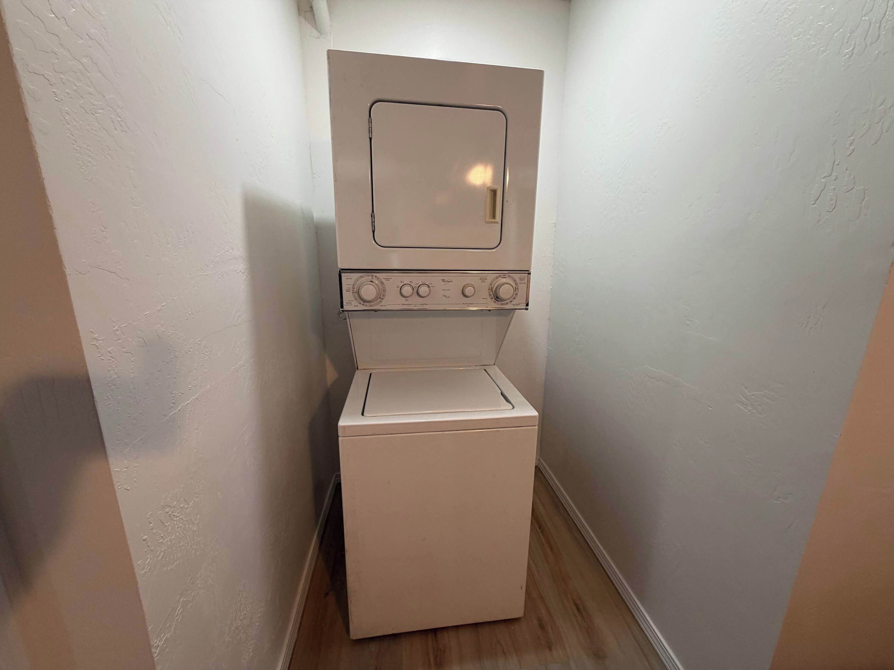 A compact, white, wall-mounted washer and dryer unit in a narrow hallway with plain walls and wooden flooring.
