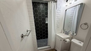 A bathroom with a white toilet, a sink with a mirror, and a shower with a black hexagonal tile wall. - 2BR Sleeps 8 Duplex | Med Ctr | Corp Stays, Family (Amarillo)
