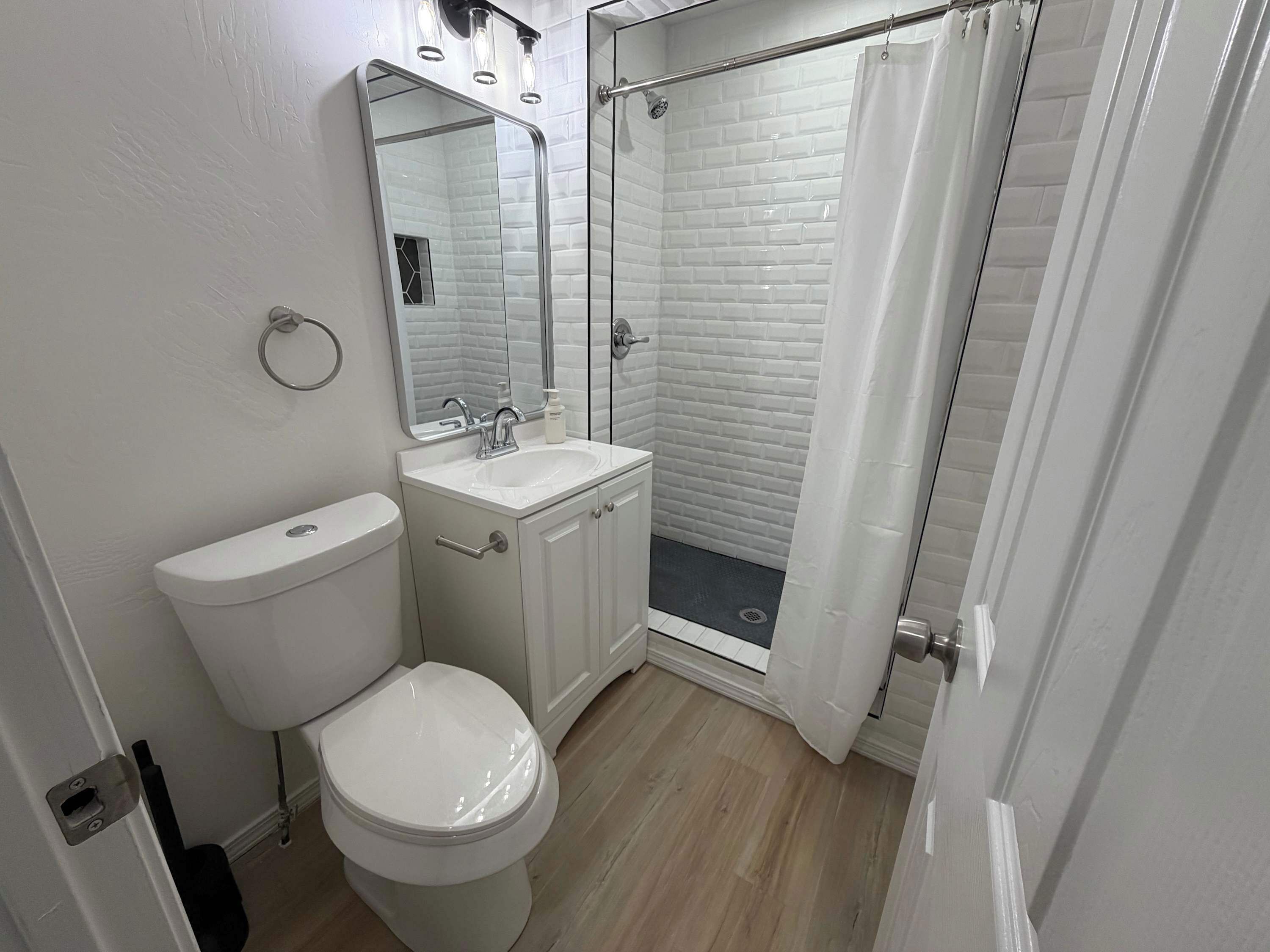 A bathroom with a toilet, sink, mirror, shower, and a door.