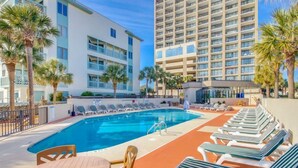 A pool area with lounge chairs, a table, and a building with balconies. - Ocean Forest Plaza 1006 (Myrtle Beach)