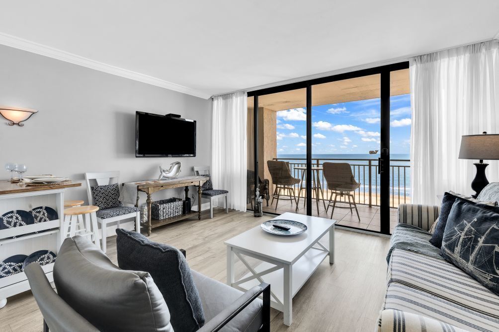 A modern living room with a flat-screen TV, a dining area, and a balcony with a view of the sea.