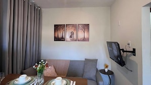 A dining area with a table set for two, a sofa, a TV mounted on the wall, and a ceiling light. - Estúdio Floripa Norte - Florianópolis-SC (Florianópolis)