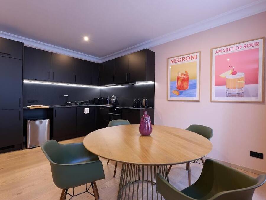 A modern kitchen with dark cabinets, a round wooden table, and green chairs. There are two framed posters on the wall.