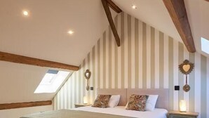 A bedroom with a sloped ceiling, a bed with a green blanket, wooden beams, a nightstand with a lamp, and striped wallpaper.