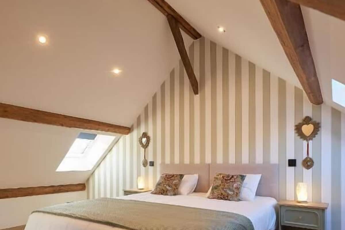 Una camera da letto con soffitto spiovente, un letto con coperta verde, travi in legno, un comodino con una lampada e carta da parati a righe.