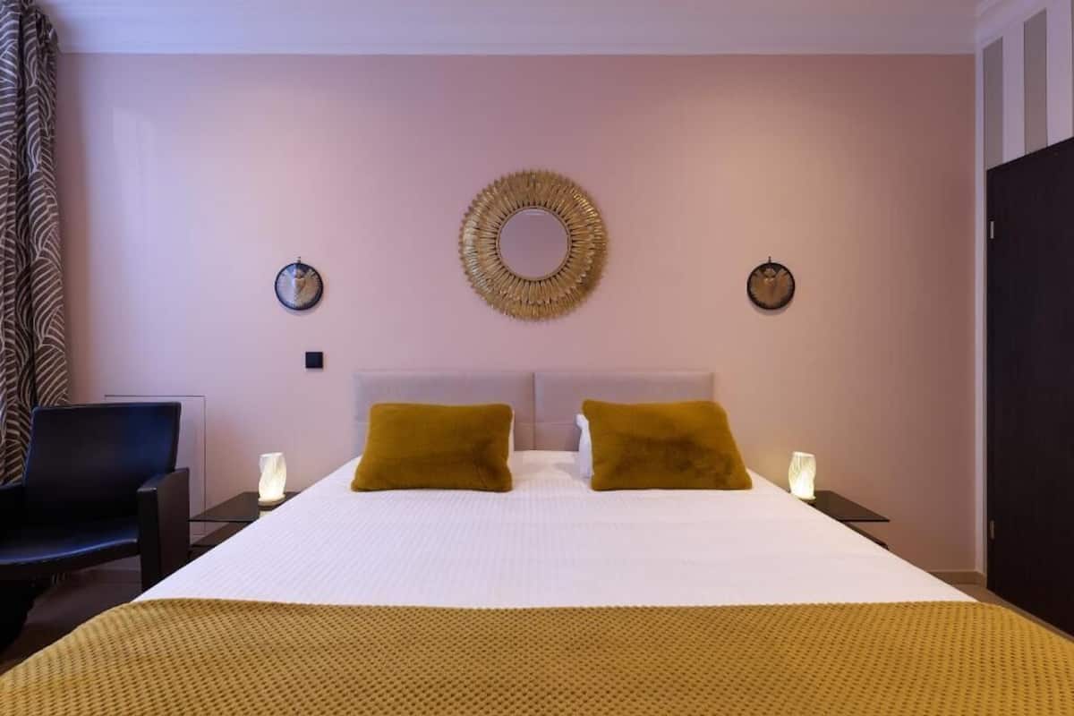 Una camera d'albergo con un letto grande, due comodini con lampade, uno specchio rotondo a parete e una sedia.