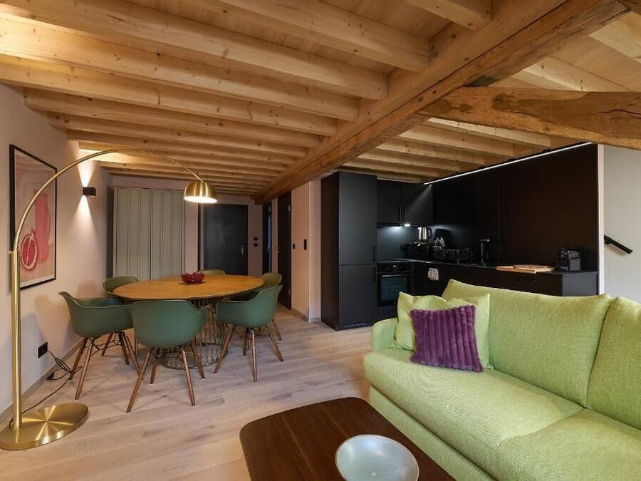 A modern living room with a wooden ceiling, a round dining table with chairs, a green sofa, and a kitchen area in the background.