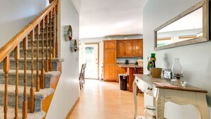 A staircase with wooden balusters, a kitchen with wooden cabinets, a dining area with chairs, and a table with a lamp and a clock.