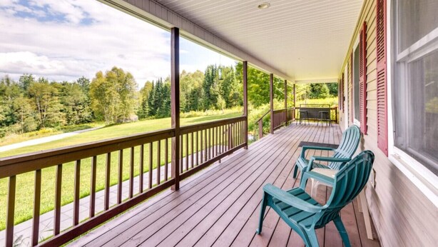 A covered porch with a view of a grassy field and trees, wooden railings, and two green chairs.