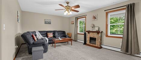 A living room with a sectional sofa, a fireplace, a ceiling fan, and large windows.