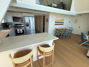 A modern kitchen with a large island, stainless steel appliances, and a dining area with wicker chairs and a wooden table. - Lux Oceanfront 4-Bed, 3-Bath Beach House. Sleeps 10. Buccaneer Beach, Oceanside (Oceanside)