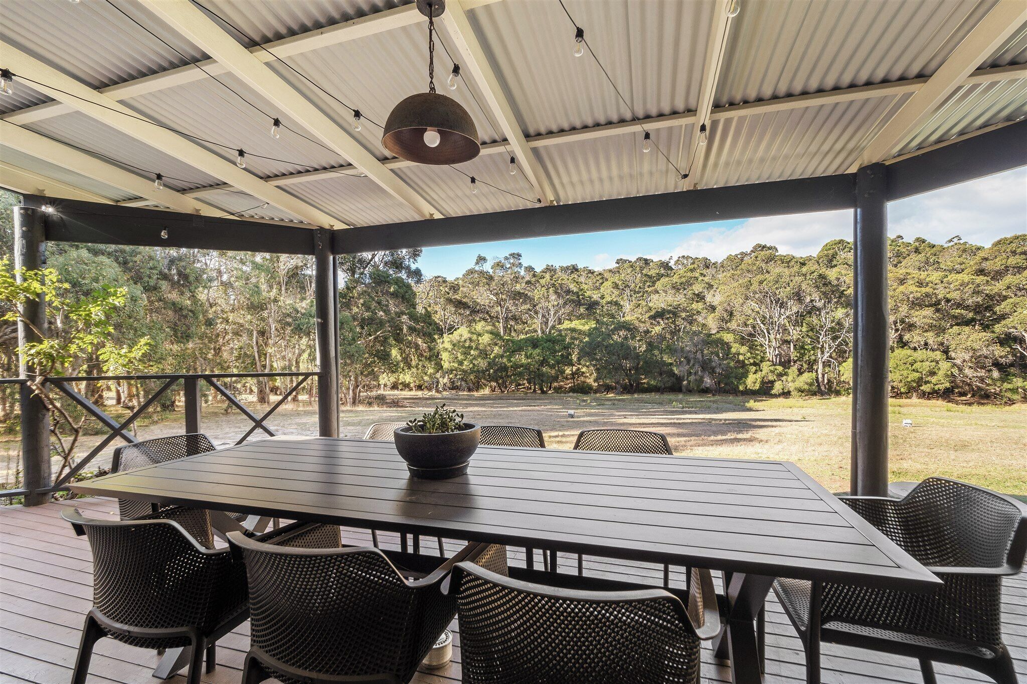 A covered outdoor area with a table and chairs, a hanging light fixture, and a view of a forested landscape.