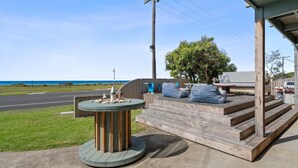 Eine Holzterrasse mit Stufen zu einem überdachten Bereich, einem runden Tisch mit einer Flagge und Blick auf das Meer.