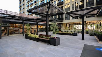 A modern outdoor area with a covered seating area, planters with greenery, and a building with multiple balconies.