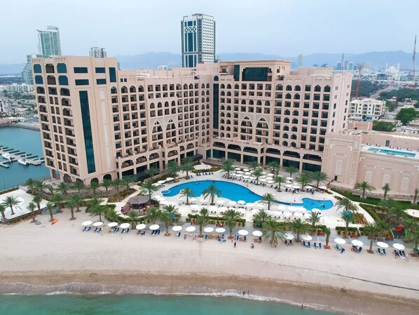 A large hotel with multiple floors, a central pool, and a beach area with sun loungers.
