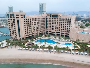 Un hotel grande con varios pisos, piscina central y zona de playa con tumbonas.