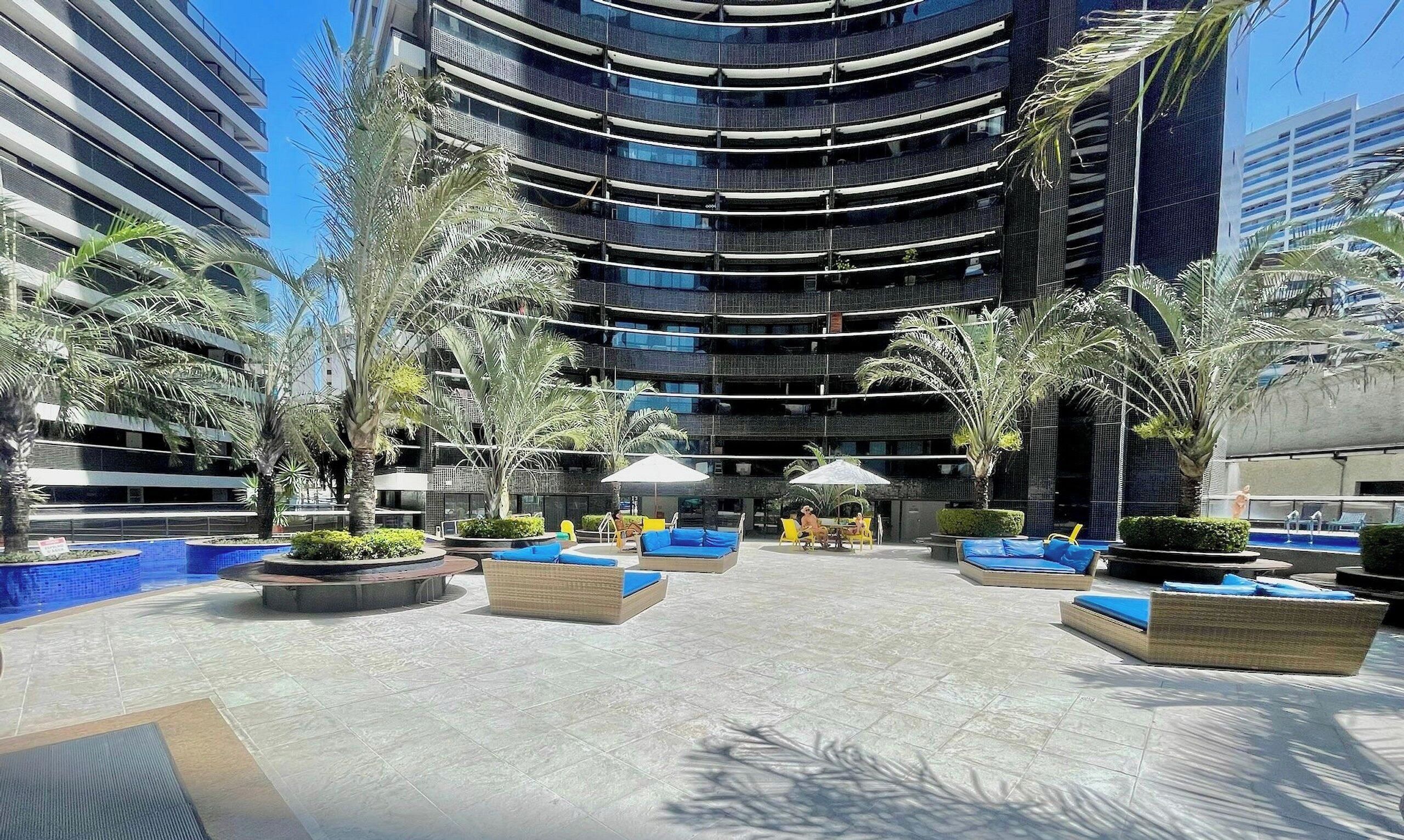 A modern hotel atrium with a central seating area, surrounded by palm trees and a pool.