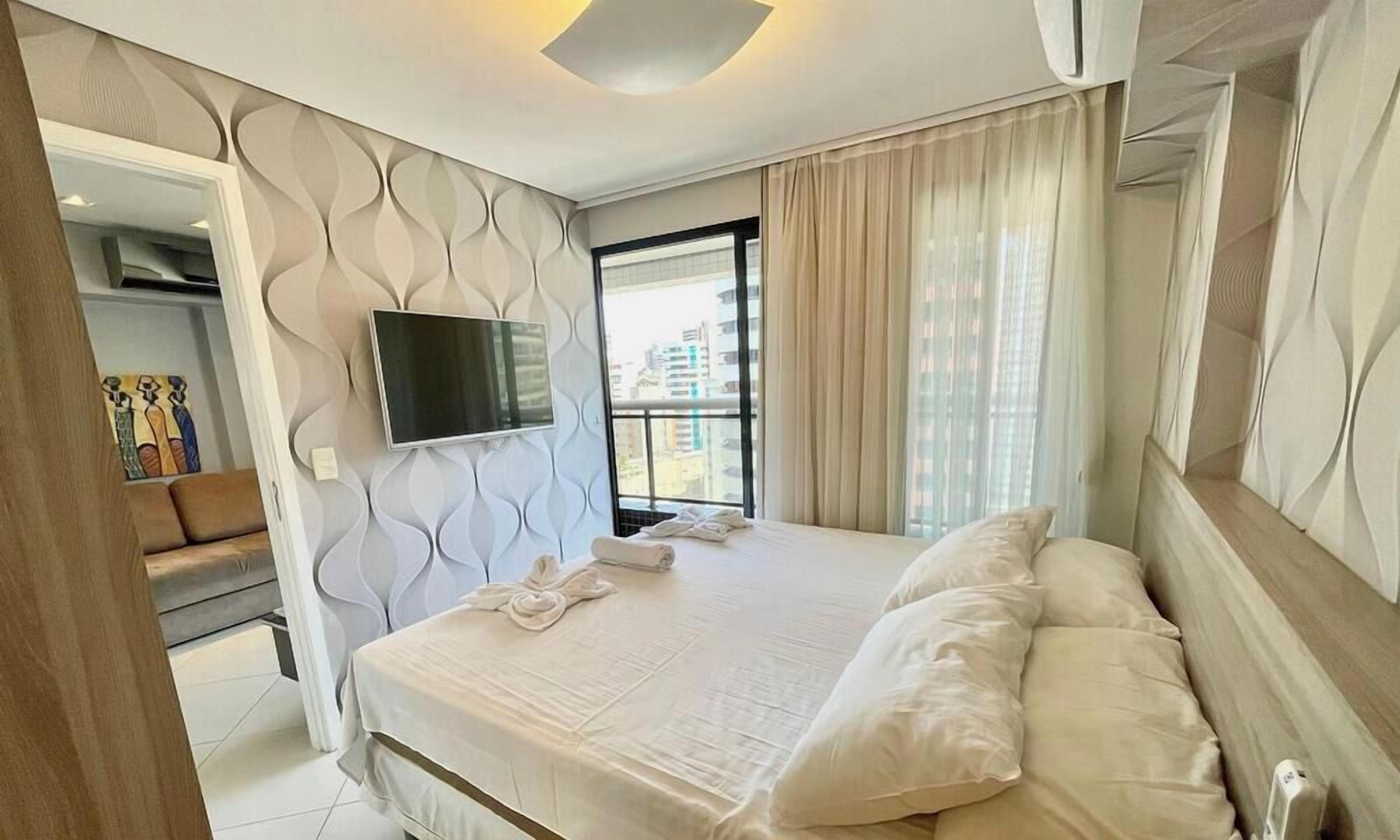 Une chambre d’hôtel avec un lit, un téléviseur fixé au mur, une fenêtre avec des rideaux et une vue sur les bâtiments environnants.