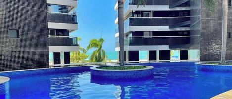 A modern hotel with a central swimming pool, multiple balconies, and a view of the ocean.