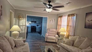 A living room with a sofa, armchair, and coffee table, a ceiling fan, and a kitchen in the background.