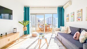 A modern living room with a sofa, a glass coffee table, a TV, and a balcony with a view of the city. - Apartment Praia Rocha | Near Beach | Safira V (Portimão)