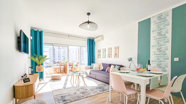 A modern living room with a sofa, dining table, and chairs, a balcony with curtains, and a wall with travel-themed decor. - Apartment Praia Rocha | Near Beach | Safira V (Portimão)