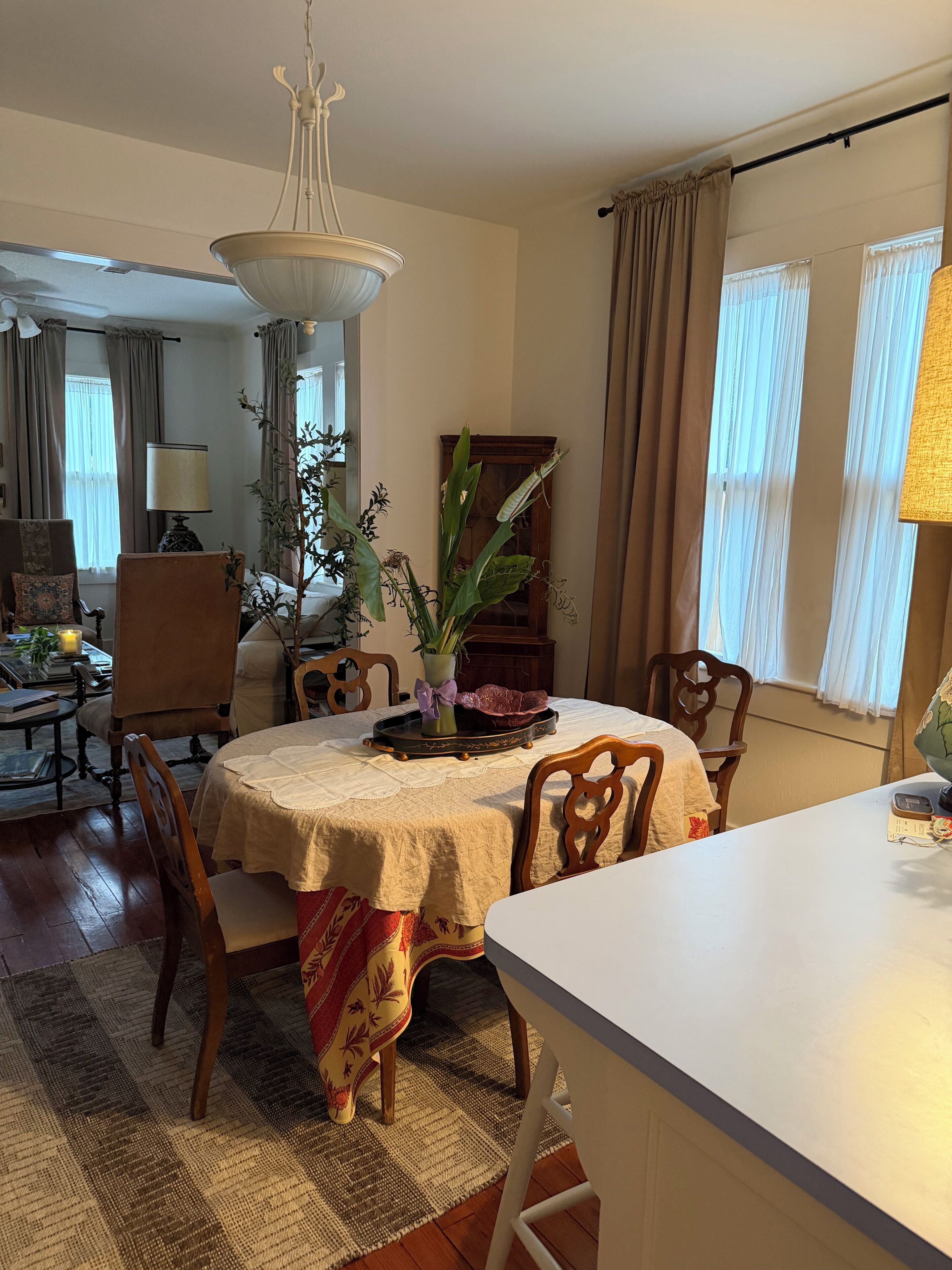 Un coin repas avec une table dressée pour quatre personnes, entourée de chaises, d’un lustre et d’une grande fenêtre avec des rideaux.