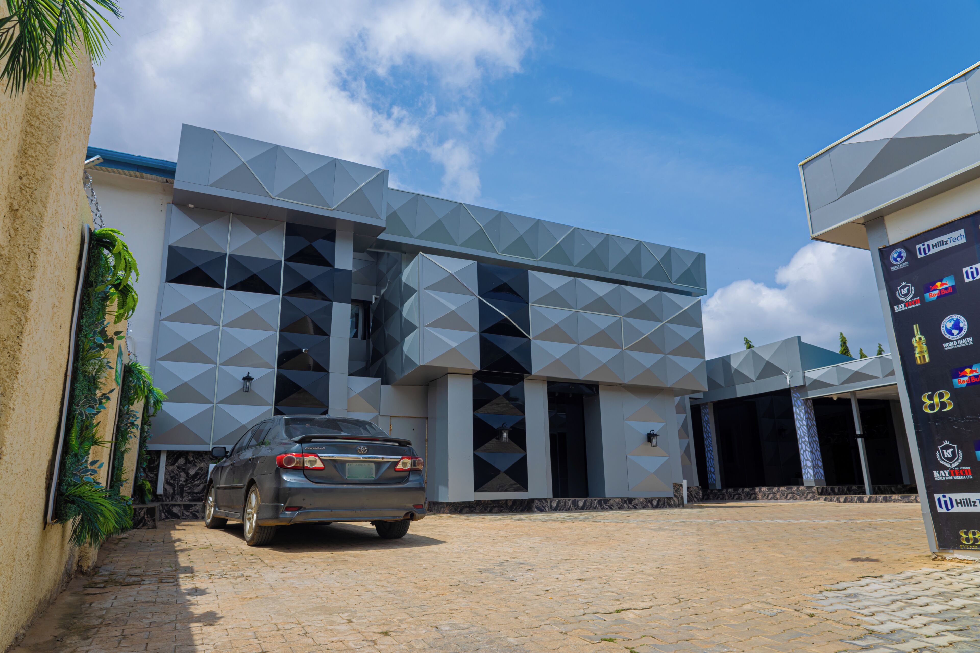 A modern building with a geometric facade, a car parked in front, and a signboard with various logos.