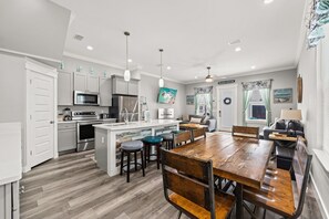 A modern kitchen with a central island, dining area with wooden chairs, and a living space with a sofa and a TV. - Rooftop Deck, Game Room, Fire Pit, Sleeps 16 (Panama City Beach)