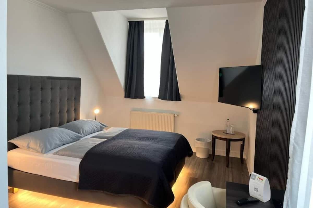 A bedroom with a bed, a TV, a small table, and a window with curtains.