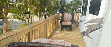 A wooden deck with a wicker chair, a lounge chair, and a white RV with a logo.