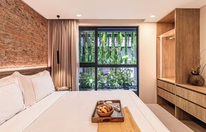 A modern bedroom with a brick wall, a large window with greenery, a bed with white linens, and a wooden bedside table with a tray of food. - Kukun Edition El Angel (Mexico City)
