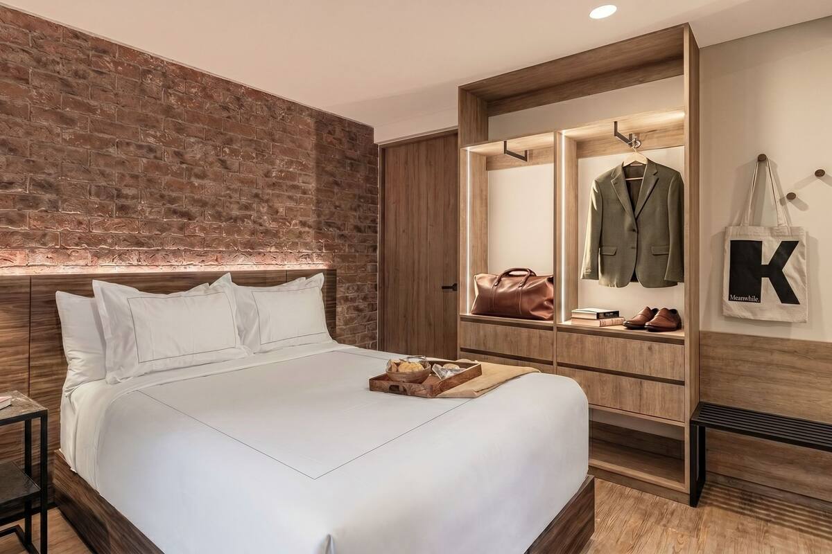 A modern bedroom with a brick wall, a wooden headboard, a bed with white linens, a bedside table, a mirror, a wardrobe, and a hanging bag with a letter 'K'.