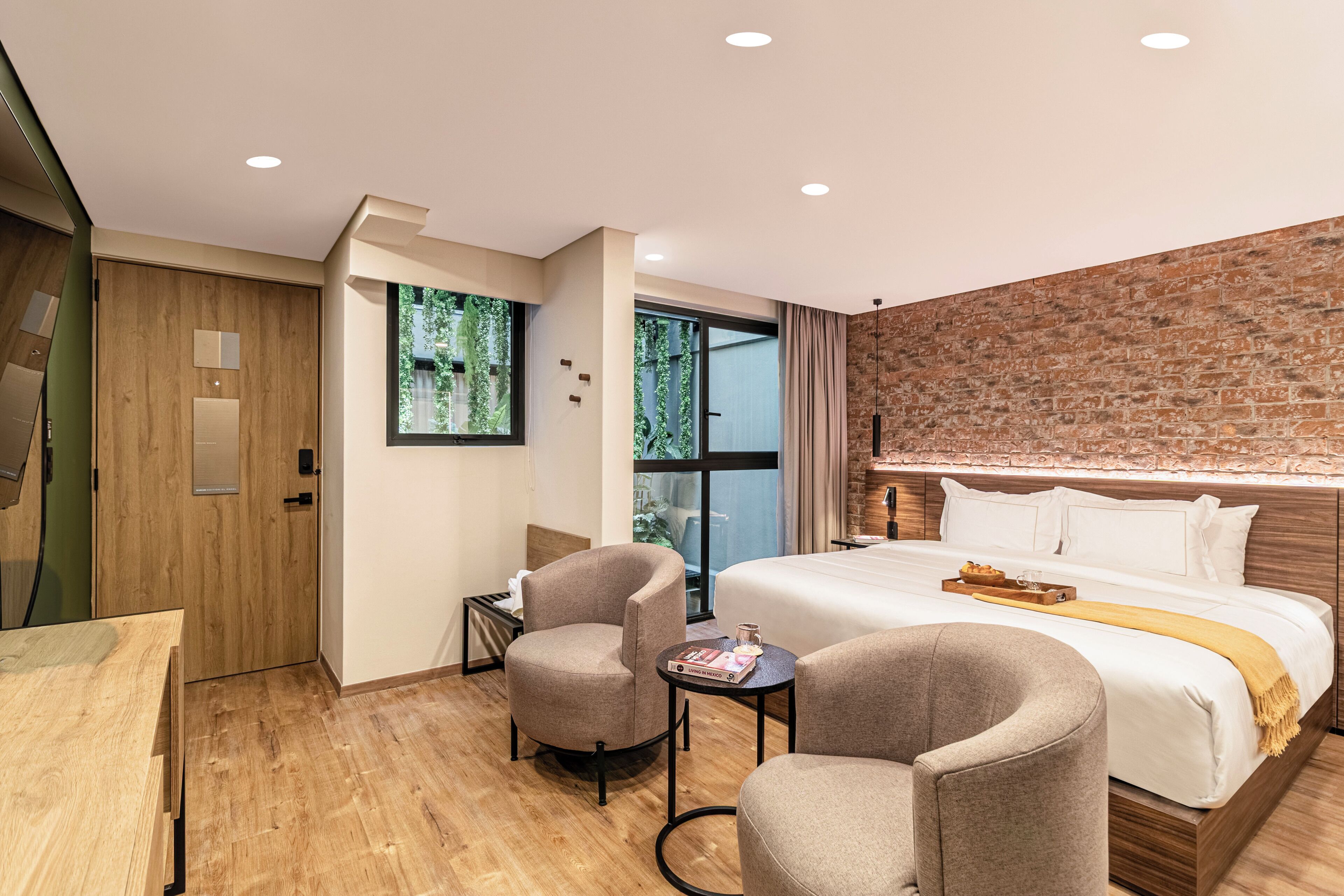 A modern hotel room with a large bed, two armchairs, a small table, a wooden wardrobe, and a brick accent wall.