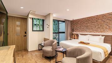 A modern hotel room with a large bed, two armchairs, a small table, a wooden wardrobe, and a brick accent wall.