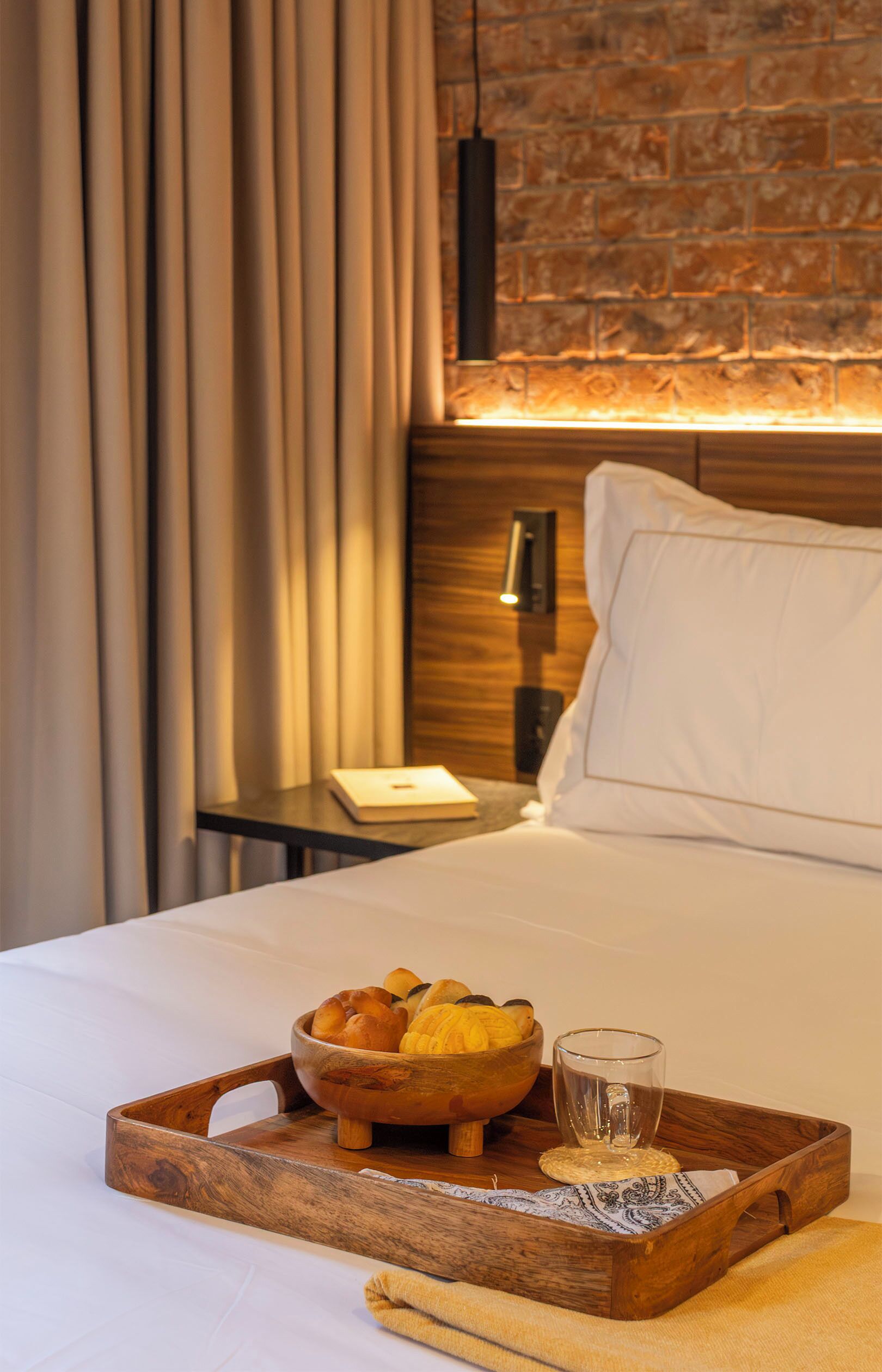 Une chambre d’hôtel avec un lit, un plateau en bois contenant du pain et un verre, et une table de chevet avec une lampe.