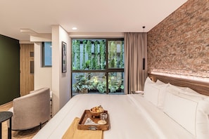 A modern hotel room with a large bed, a wooden side table with breakfast items, a chair, and a view of greenery through a large window.