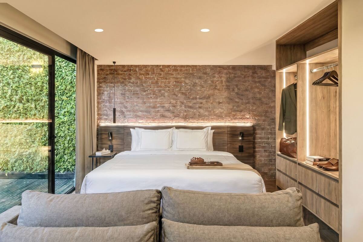 A modern hotel room with a large bed, a wooden headboard, a built-in wardrobe, and a sliding glass door to a balcony.