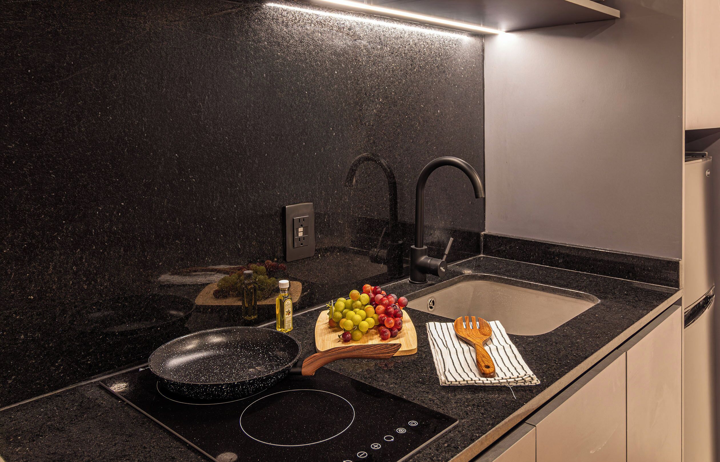 Una cocina con encimera negra, un lavabo, un grifo, una sartén, tabla de cortar con fruta y una botella de aceite.
