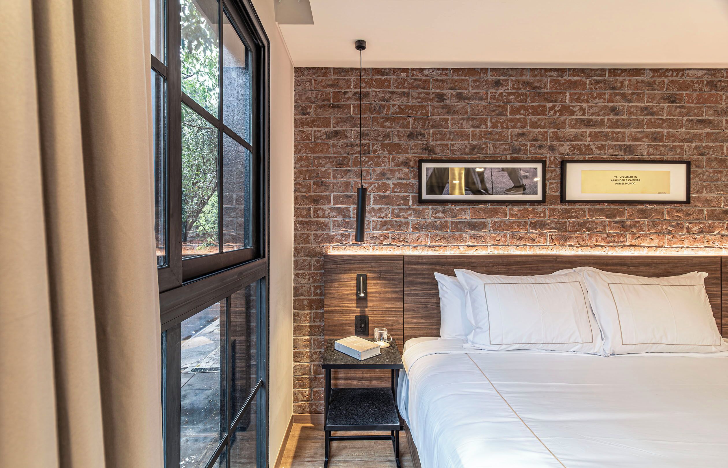 A hotel room with a brick wall, a bed with white linens, a nightstand with a book and a glass, and framed pictures on the wall.
