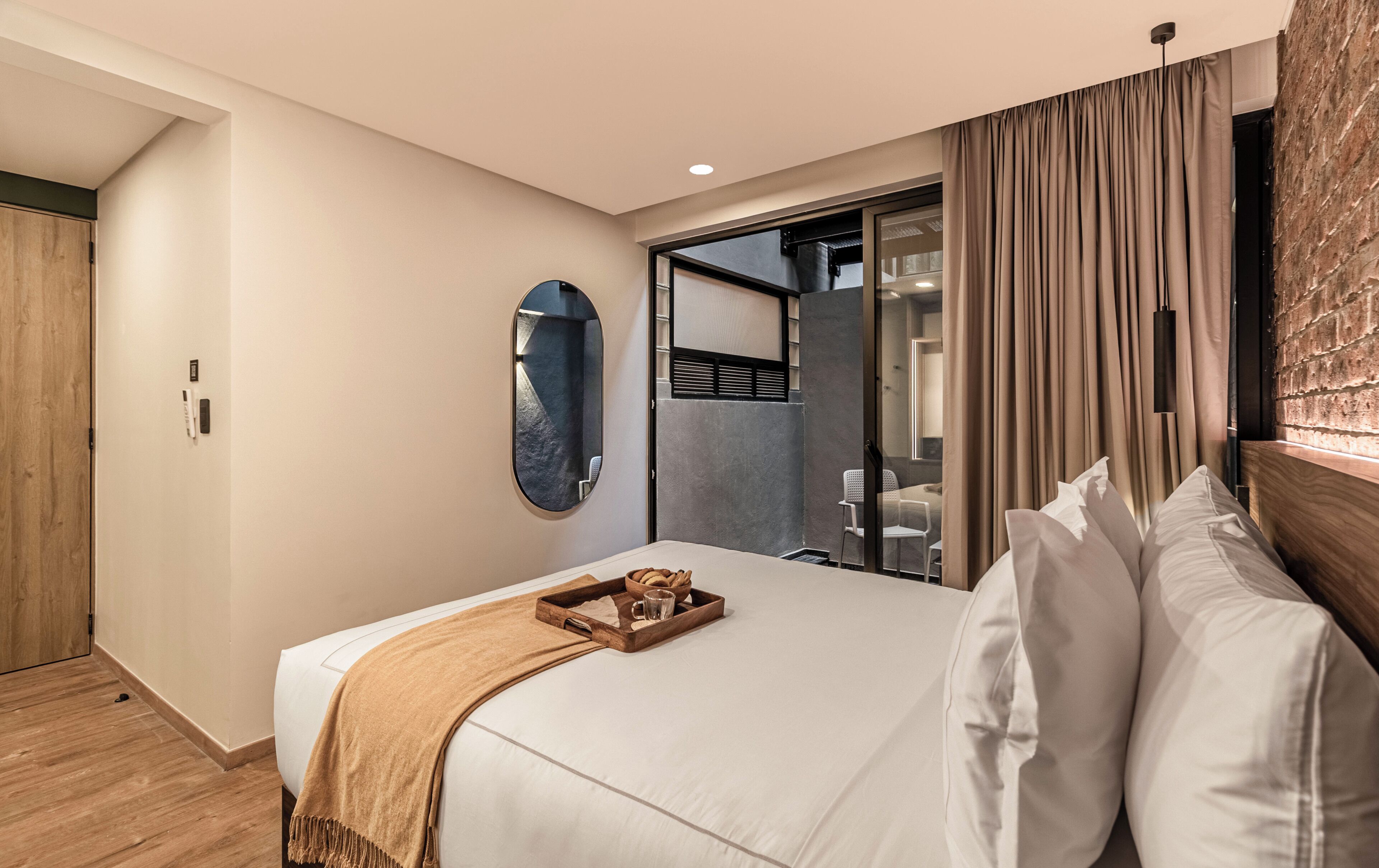 A hotel room with a bed, a wooden side table, a mirror, and a sliding glass door leading to a bathroom.