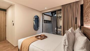 A hotel room with a bed, a wooden side table, a mirror, and a sliding glass door leading to a bathroom.