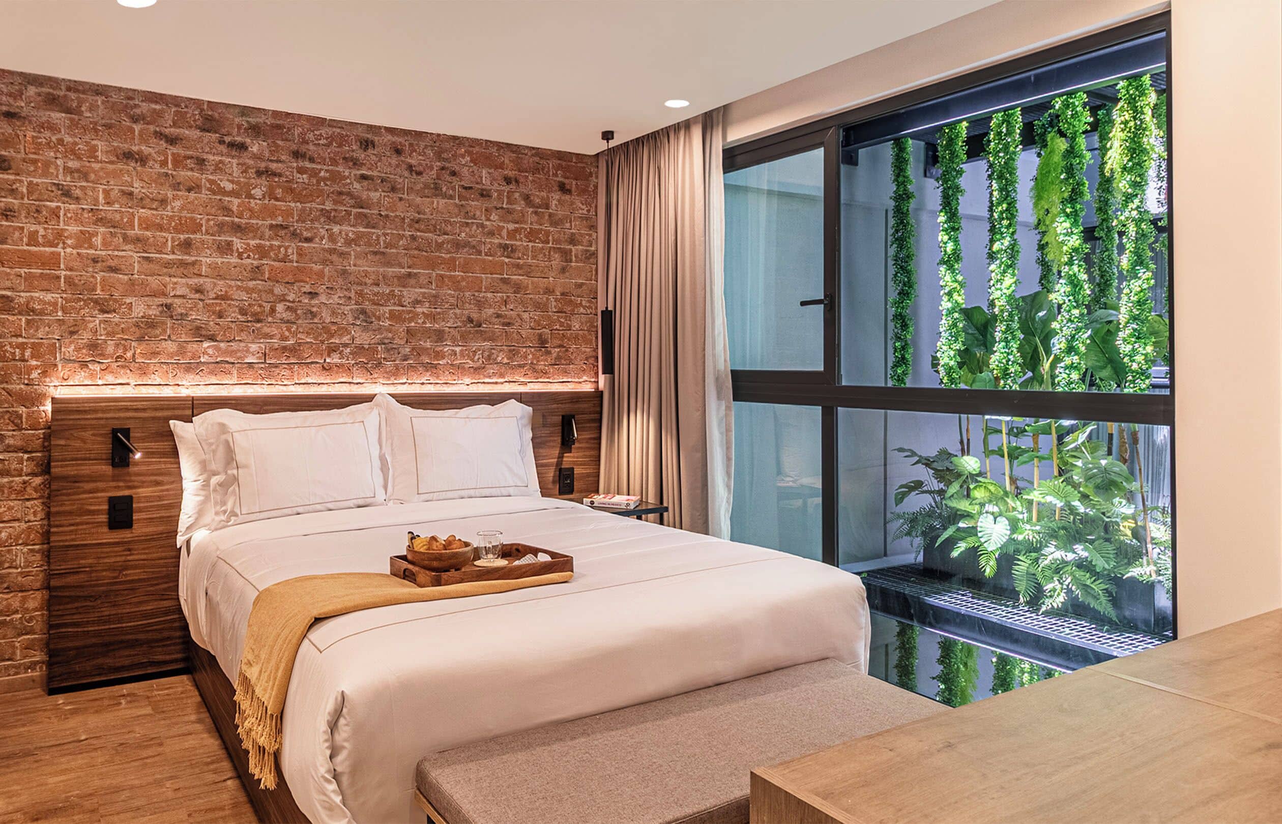 A hotel room with a brick wall, a large window with a view of greenery, a neatly made bed, and a breakfast tray on the side.