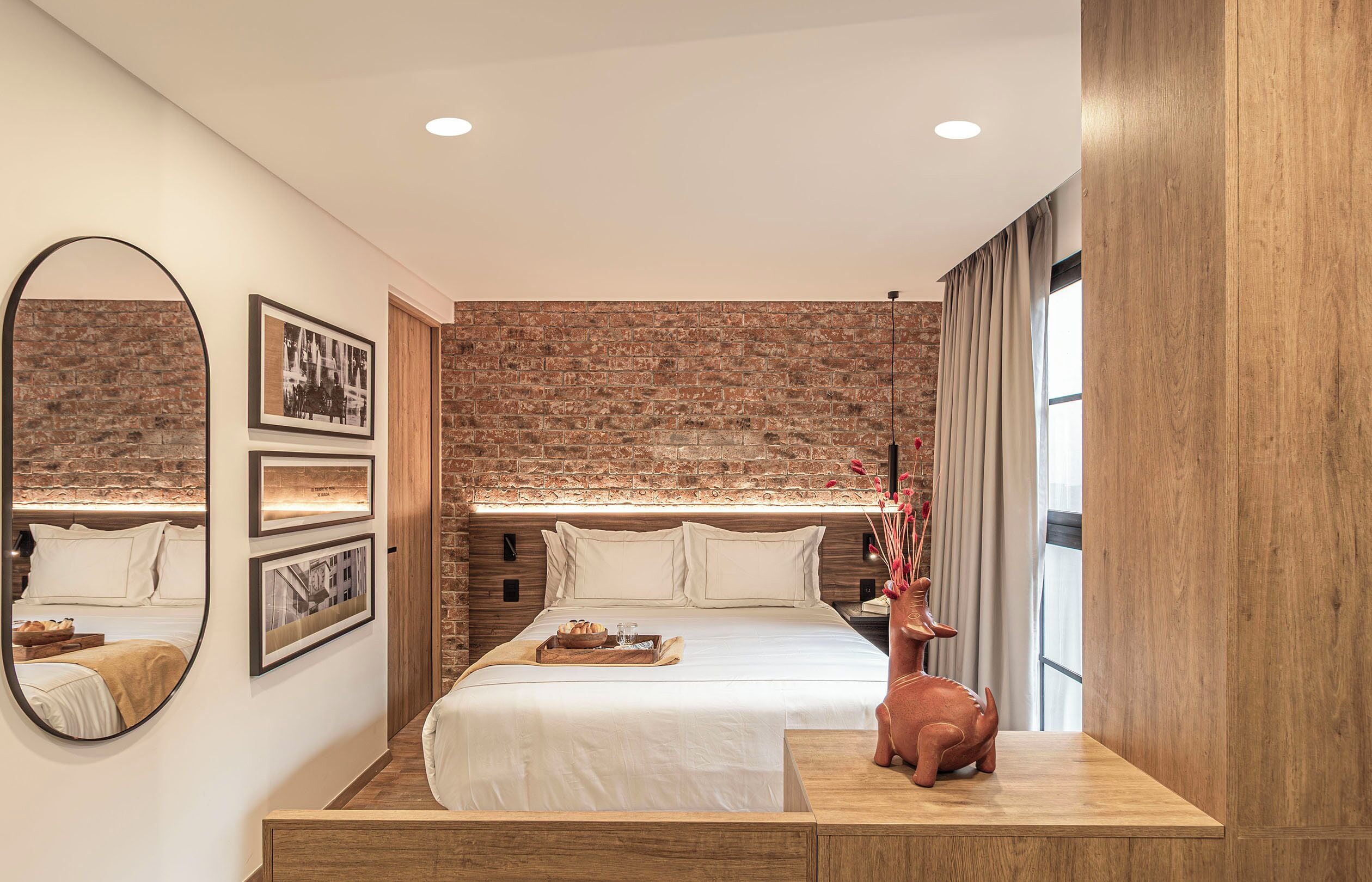 Une chambre d’hôtel avec un mur en briques, une tête de lit en bois, un lit avec du linge de maison blanc, une table de chevet, un miroir et des tableaux encadrés accrochés au mur.