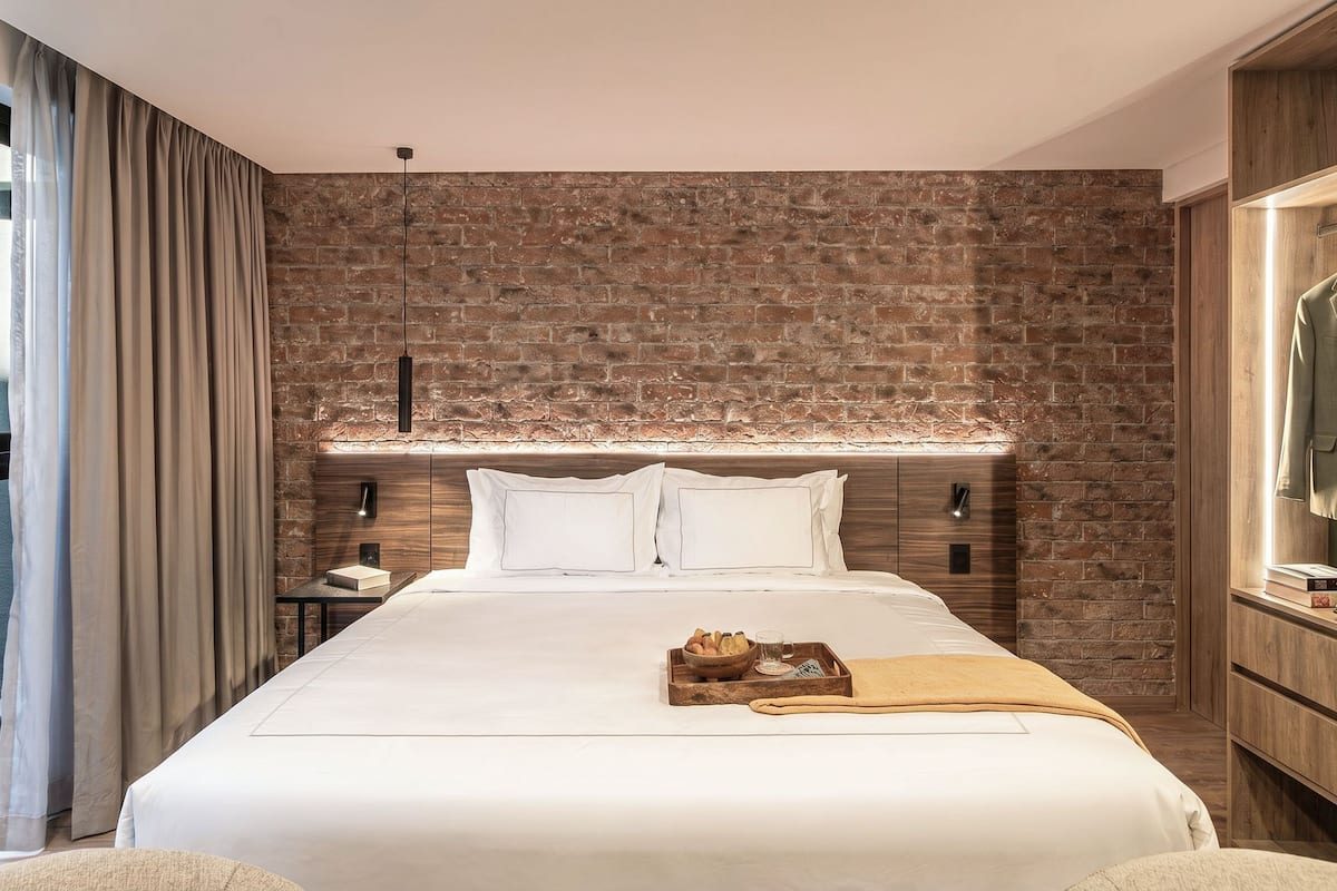 A hotel room with a brick wall, a bed with white bedding, a wooden nightstand, and a TV on the right.
