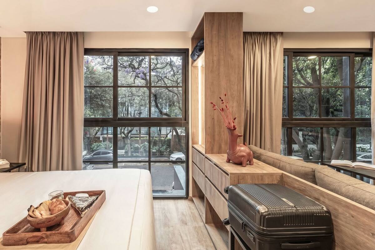 A hotel room with a bed, a desk, a suitcase, and large windows with a view of trees.